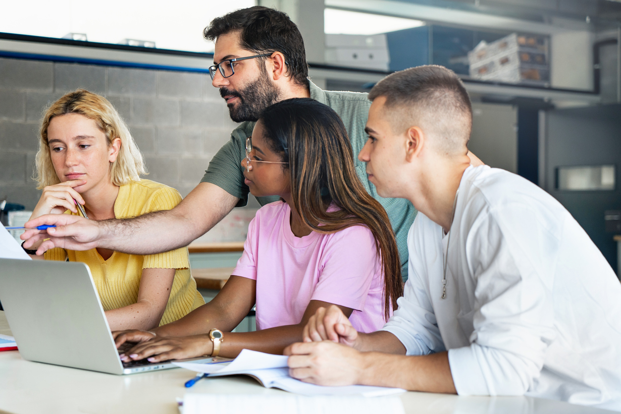 A high school teacher helps three students in a classroom.
