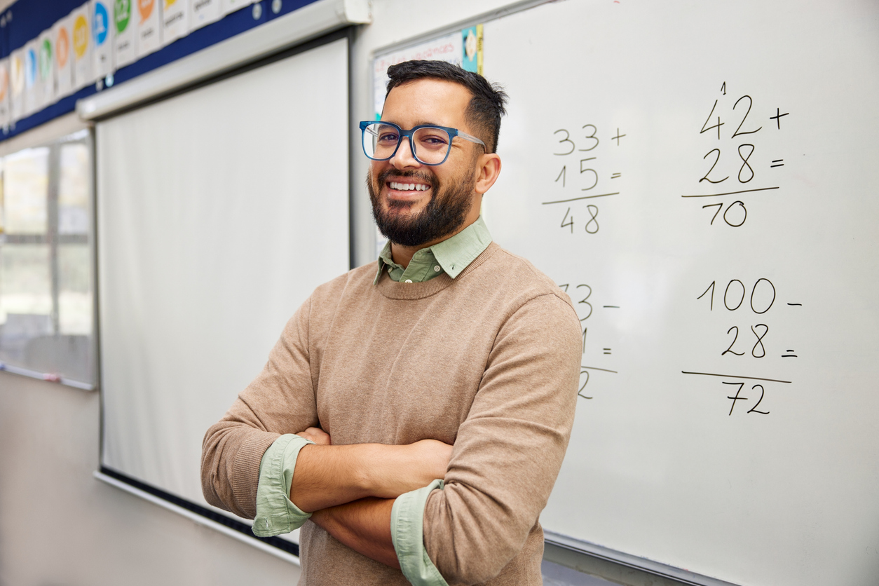 A Smiling Teacher Stands in a Classroom in Front of a Dry Erase Board