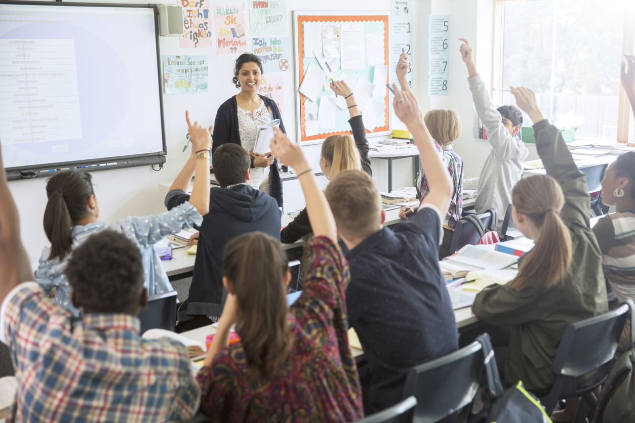 An ESL teacher stands in front of students raising their hands in a classroom.