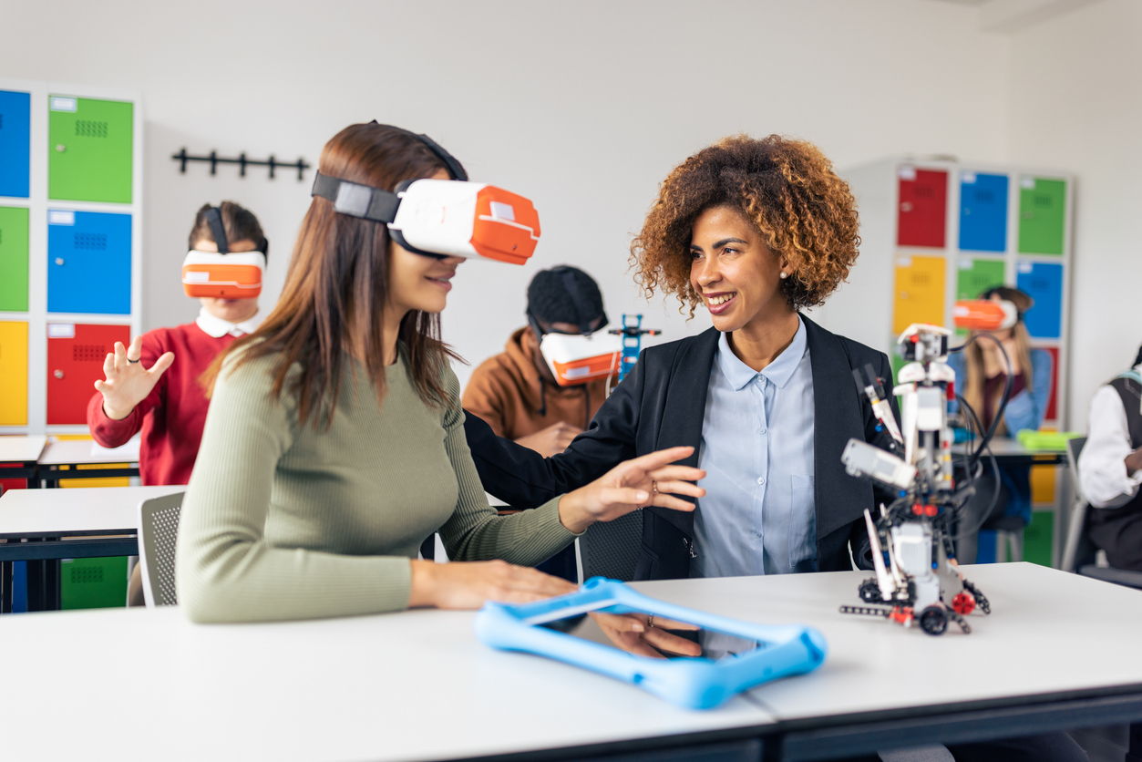 A Student Uses a Vr Headset to Interact With a Robot While a Smiling Teacher Observes.