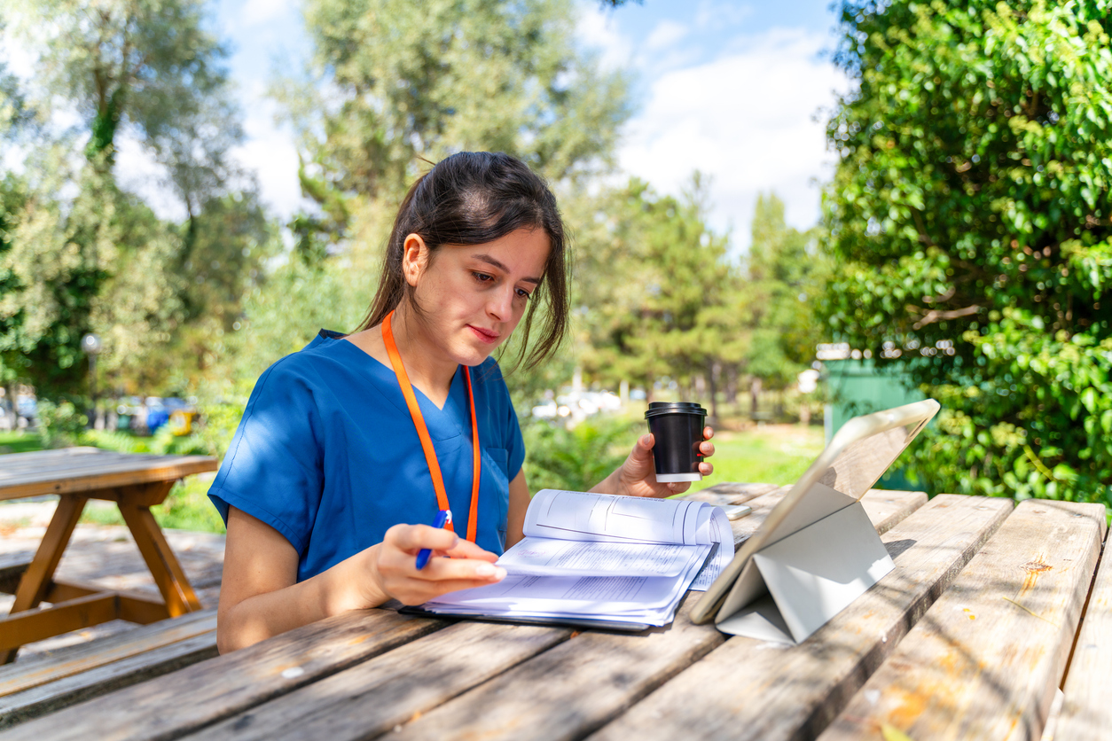 A nursing student with paperwork and a tablet studies outside at a park bench.