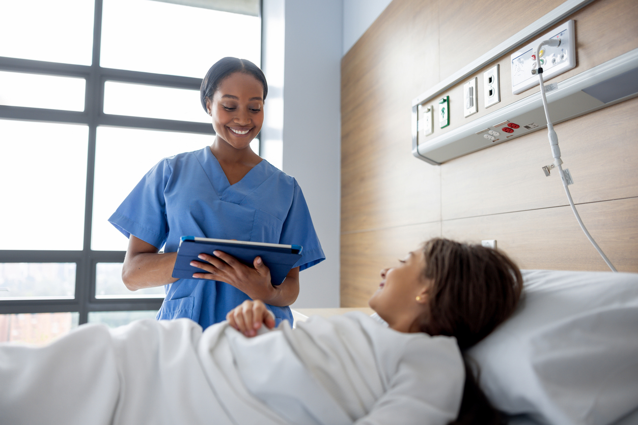 A Nurse Holding a Tablet Stands Next to a Patient in a Hospital Bed.