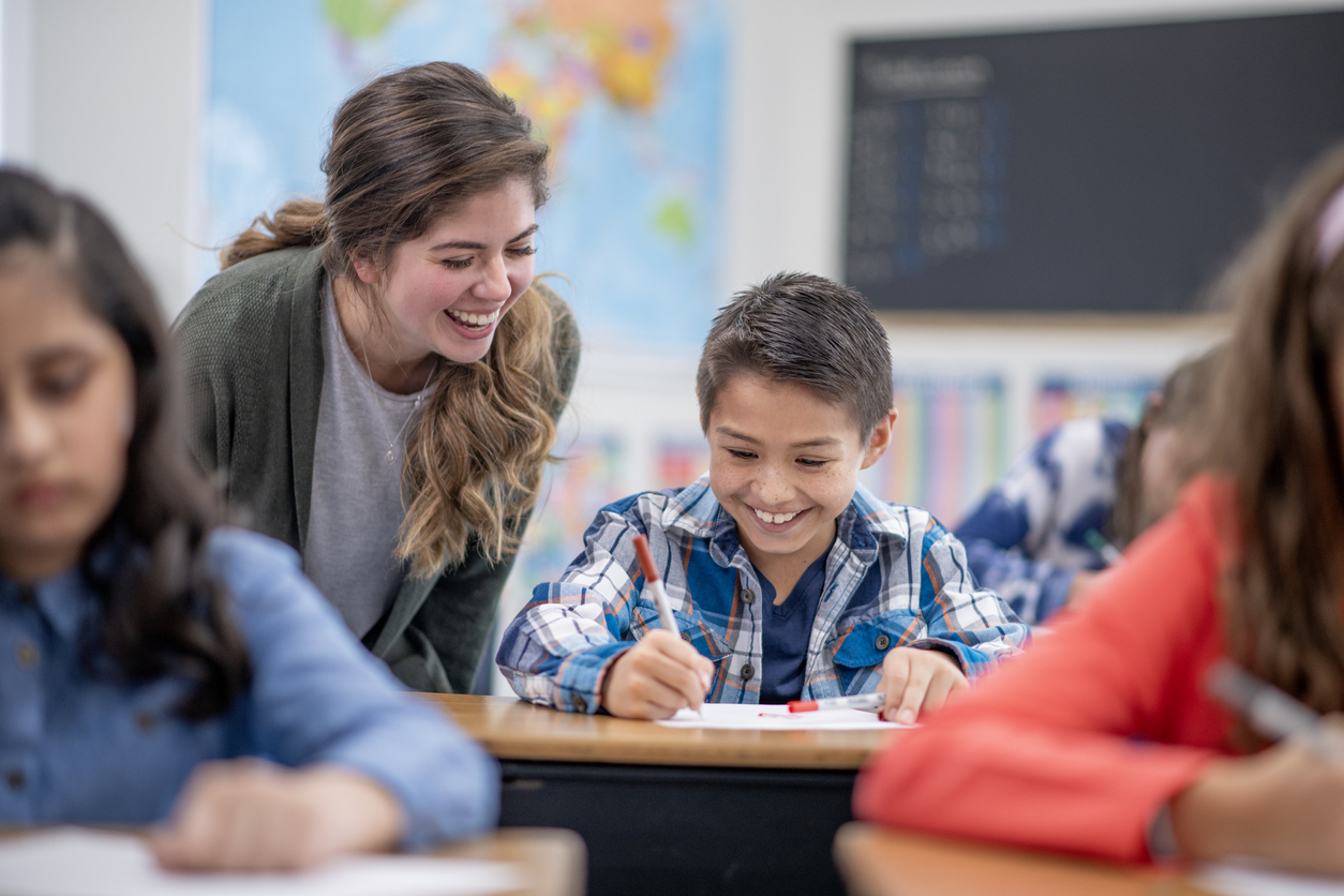 A Smiling Teacher Helps a Smiling Student Who Is Sitting at a Desk and Writing.