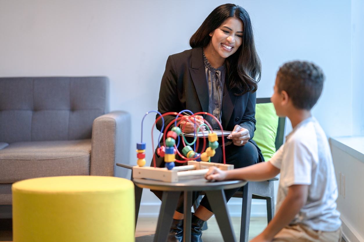 A child development specialist holding a clipboard and interacting with a child playing with a toy on a table.