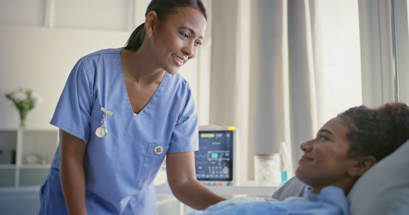 A Smiling Nurse Practitioner Speaks to a Patient Who Is Laying in a Hospital Bed
