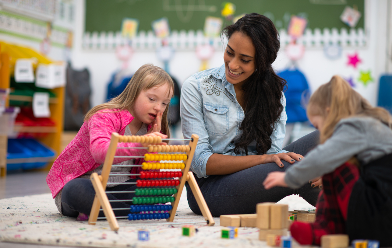 A Special Education Teacher Sits on the Floor With Two Students in a Classroom.