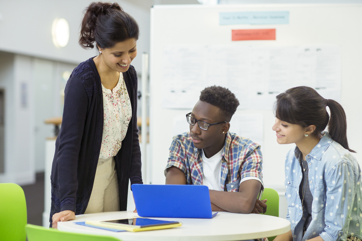 A Tesol Teacher Standing Next to Two Students Who Are Working on a Laptop.