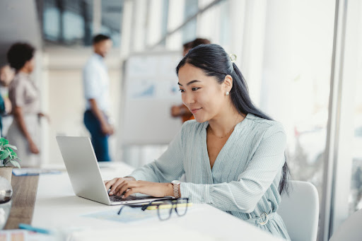An employee works on a laptop while other members of the team meet in the background.