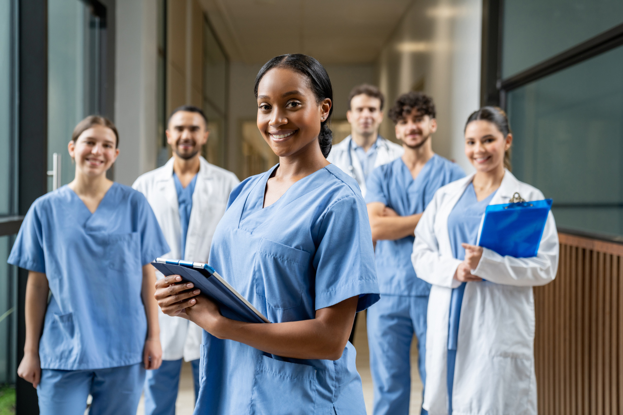 A Smiling Nurse Stands in Front of a Group of Nurses and Medical Professionals in a Hospital Hallway.