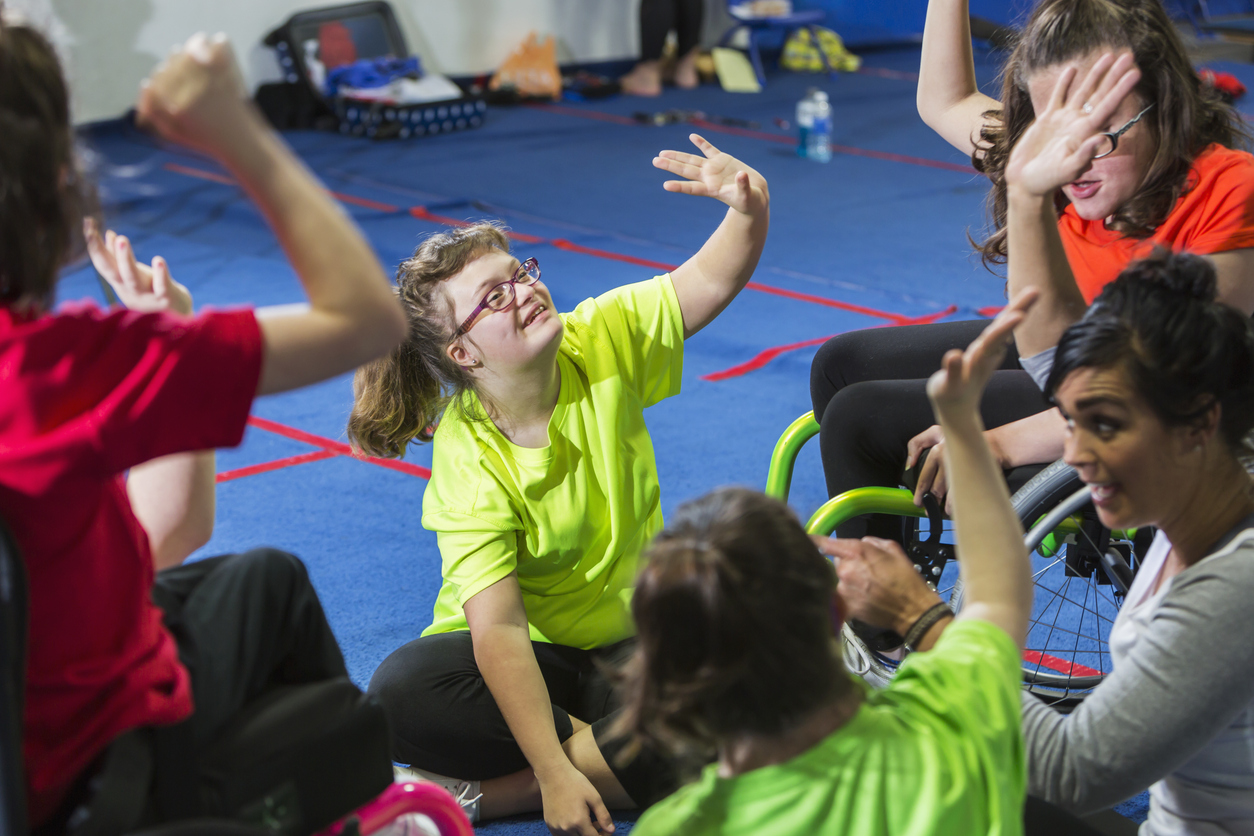 A Group of Students in an Adaptive Physical Education Class Sit on an Exercise Mat and Raise Their Hands